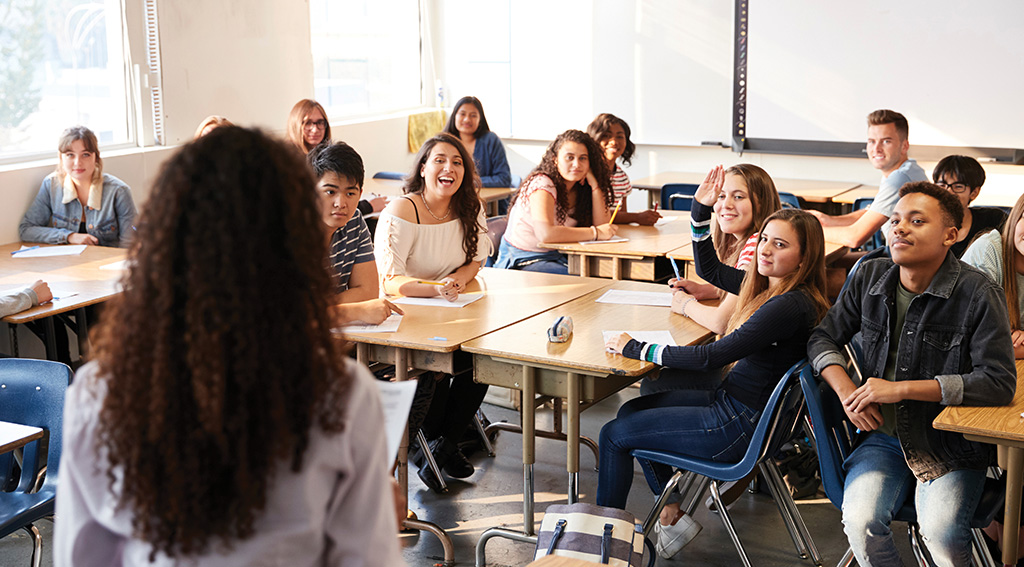 A group of students sitting at desks focus on a teacher in the front of the classroom. 