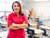 A teacher stands with her arms folded across her chest in an empty classroom. 