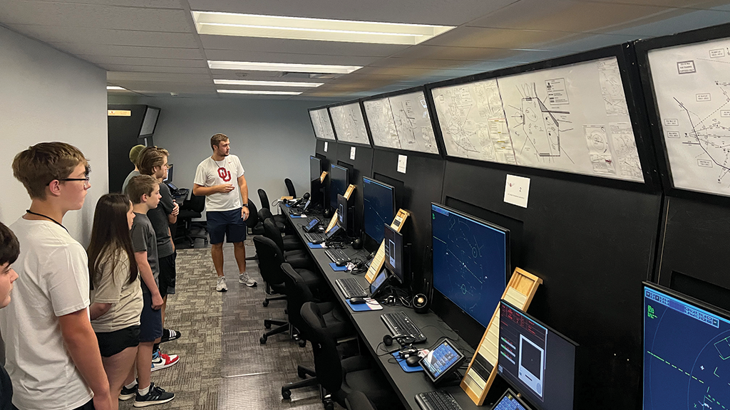 A group of high school students listen to a tour guide showing them the computers and screens in an air traffic control simulator lab,