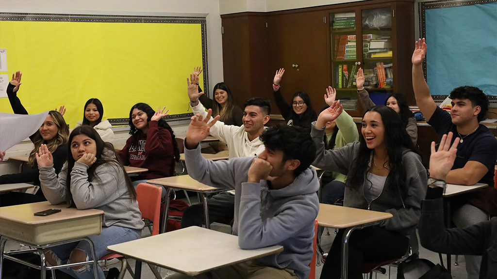 high school students sitting at desks eagerly raise their hands to answer a question