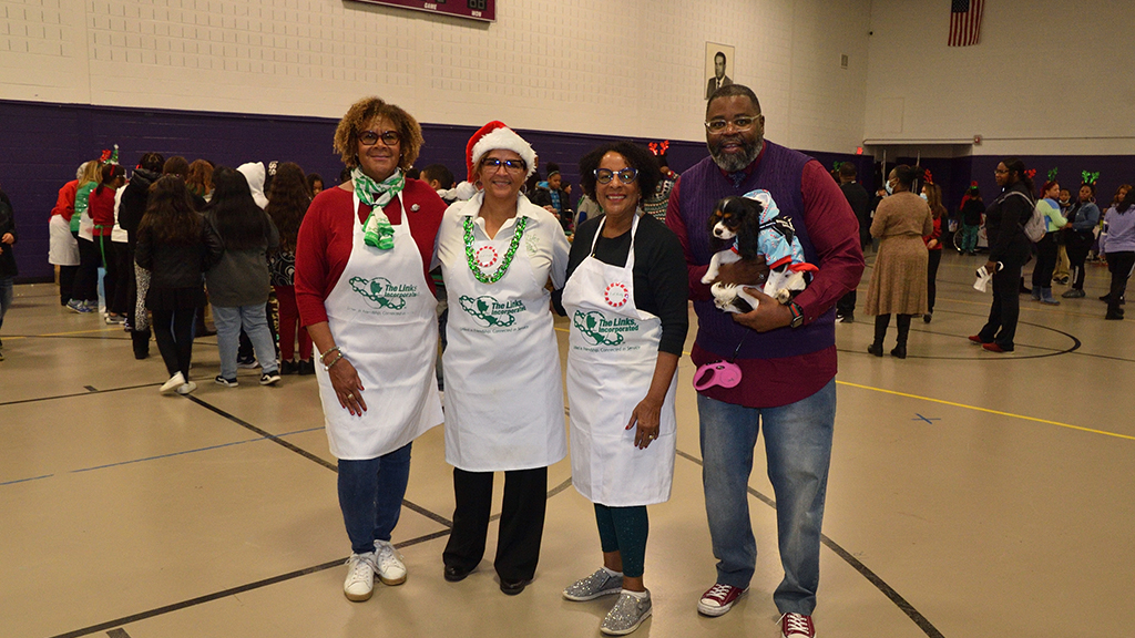 Three women wearing aprons stand next to a man holding a small dog in his arms during a holiday party