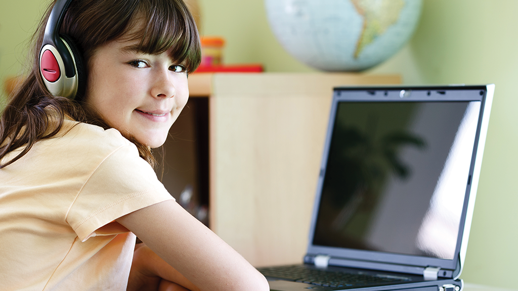a girl with headphones on sits in front of a computer screen and smiles at the camera