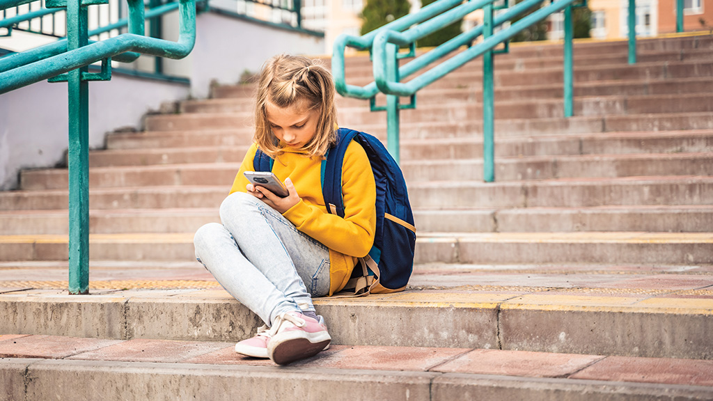 An elementary-age girls sits on her school steps engrossed in the activity shown on her phone. 
