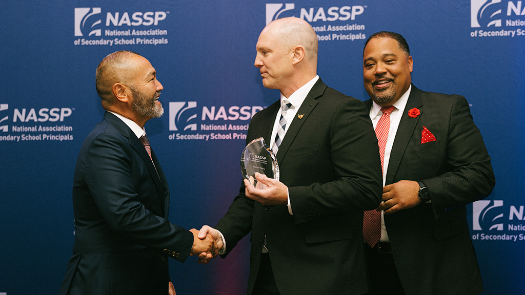 A man in a suit holding a trophy shakes hands with another man while a third man standing the background smiles.