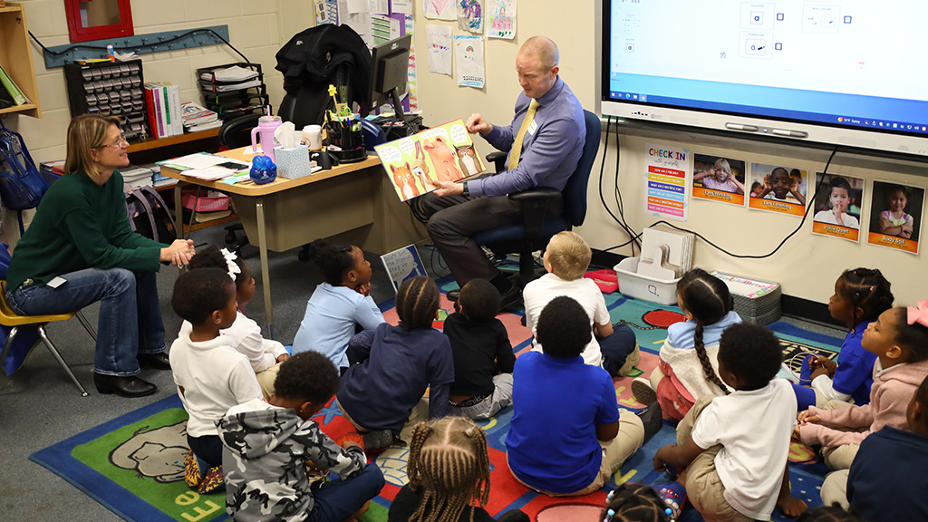 A man sits in a chair and reads a story to a group of young children sitting on the floor. 