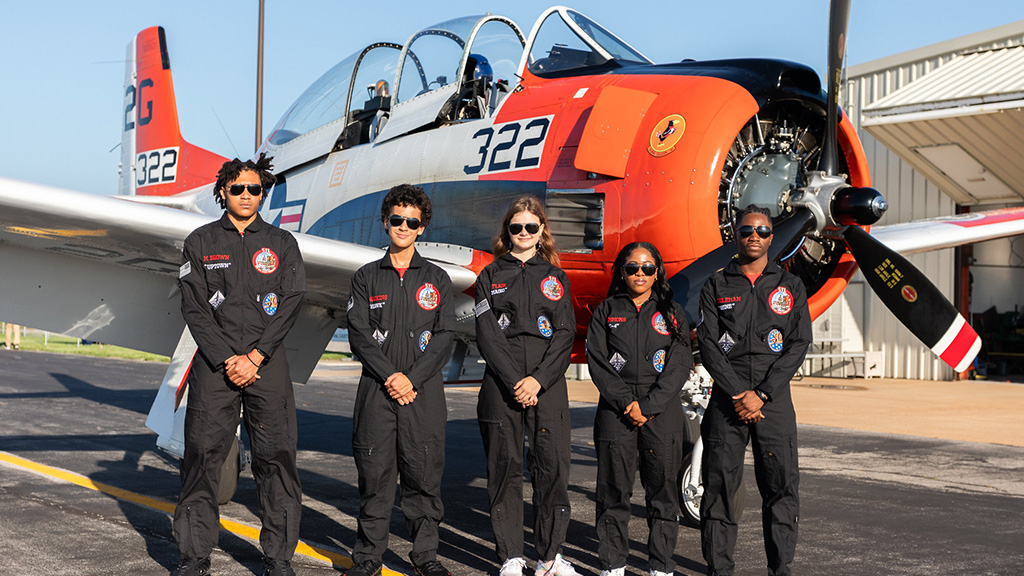 A group of five student aviators dressed in flight suits and wearing sunglasses stand in front of an airplane. 
